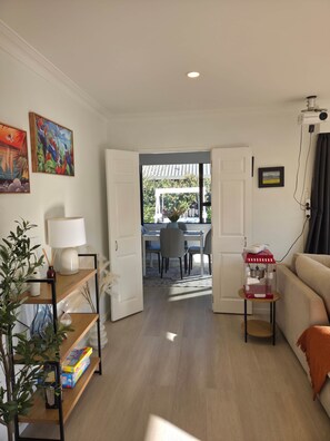 A living room with a sofa, a coffee table, a lamp, and a bookshelf. There is a dining area visible through an open door.