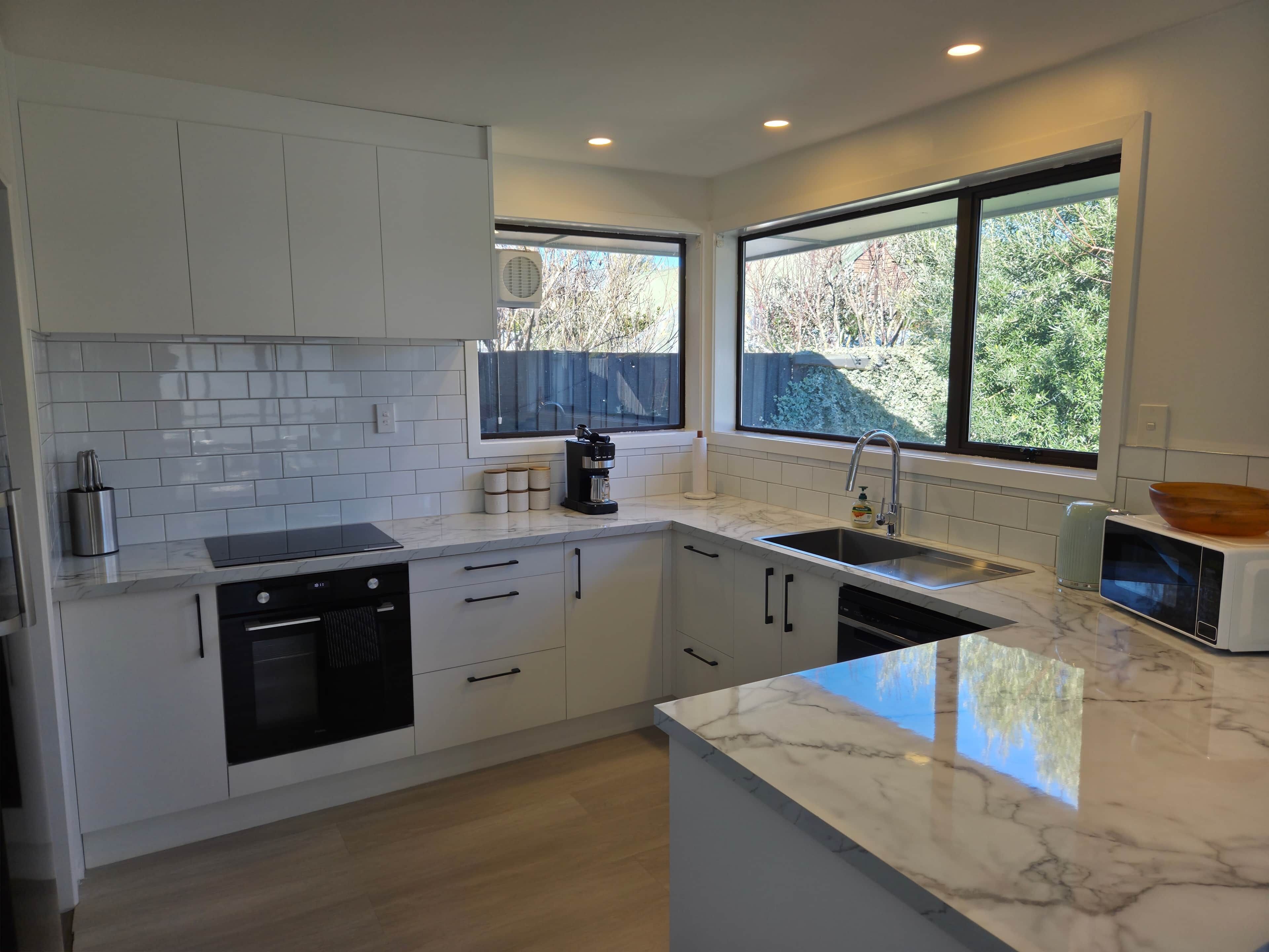 A modern kitchen with white cabinets, a marble countertop, a built-in dishwasher, and a window with a view of greenery.