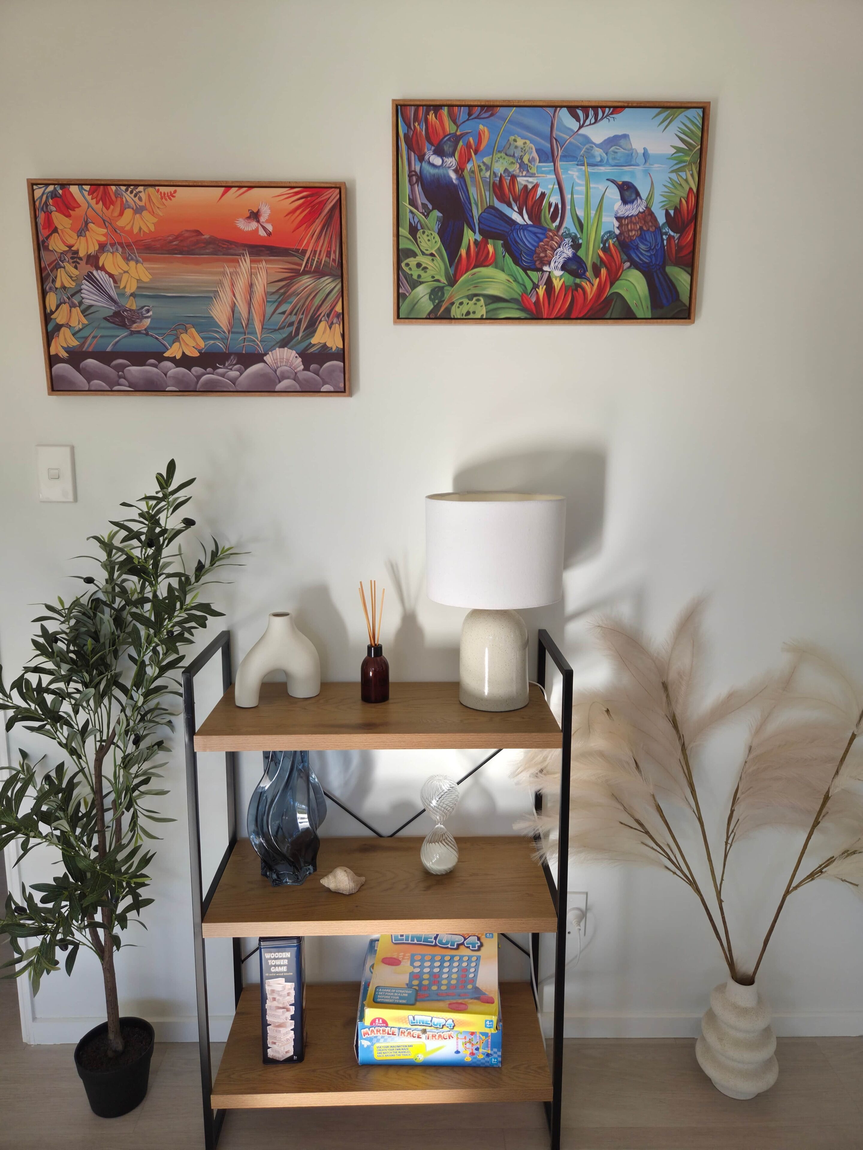 Um quarto com uma estante de madeira com uma lâmpada, uma planta e um jogo de corrida de mármore. Há duas pinturas na parede.