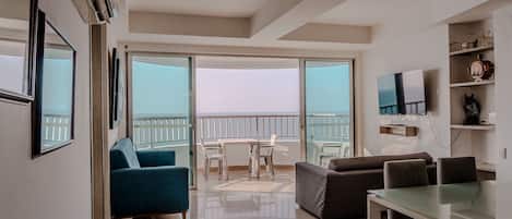 Una sala de estar moderna con un ventanal que da a un balcón, un comedor y un televisor montado en la pared.