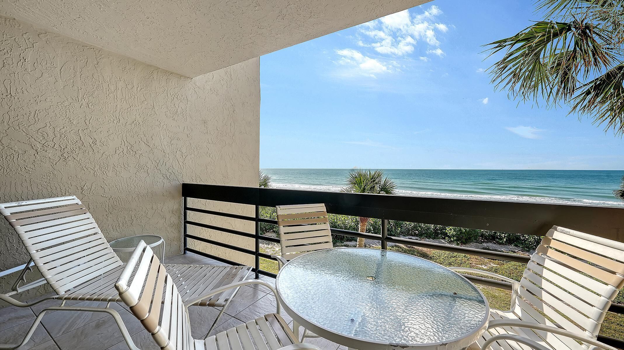 Een balkon met een glazen tafel en witte stoelen, met uitzicht op een strand met helderblauw water en een palmboom.
