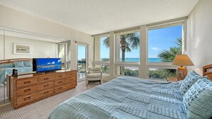 Ein Schlafzimmer mit einem großen Bett, einem Fernseher auf einer Holzkommode und Blick auf das Meer durch große Fenster.