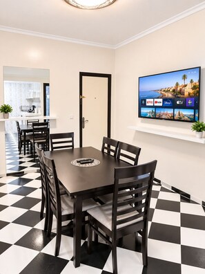 Un comedor con piso a cuadros, una mesa de madera oscura y sillas. Un televisor montado en la pared muestra varios servicios de streaming.