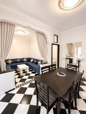 A modern living room with a black and white checkered floor, a dark wooden dining table with chairs, a blue sofa with cushions, and a white coffee table. - Large Studio Fiber optics IPTV (Marrakech)
