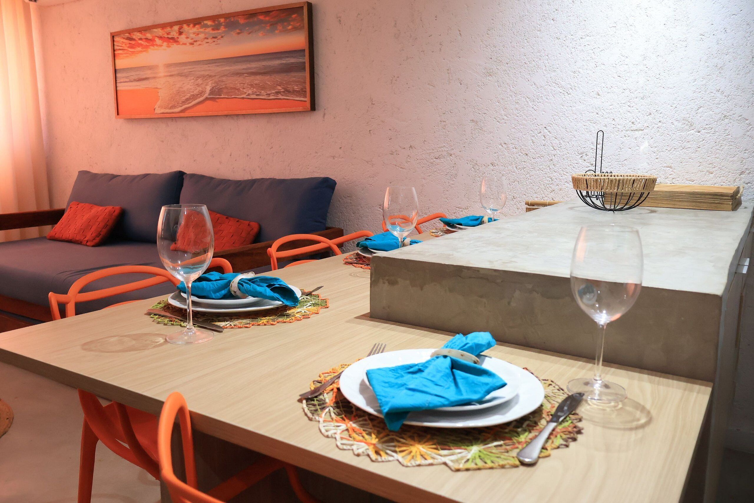A dining area with a table set for two, a sofa with cushions, a painting on the wall, and a concrete block.