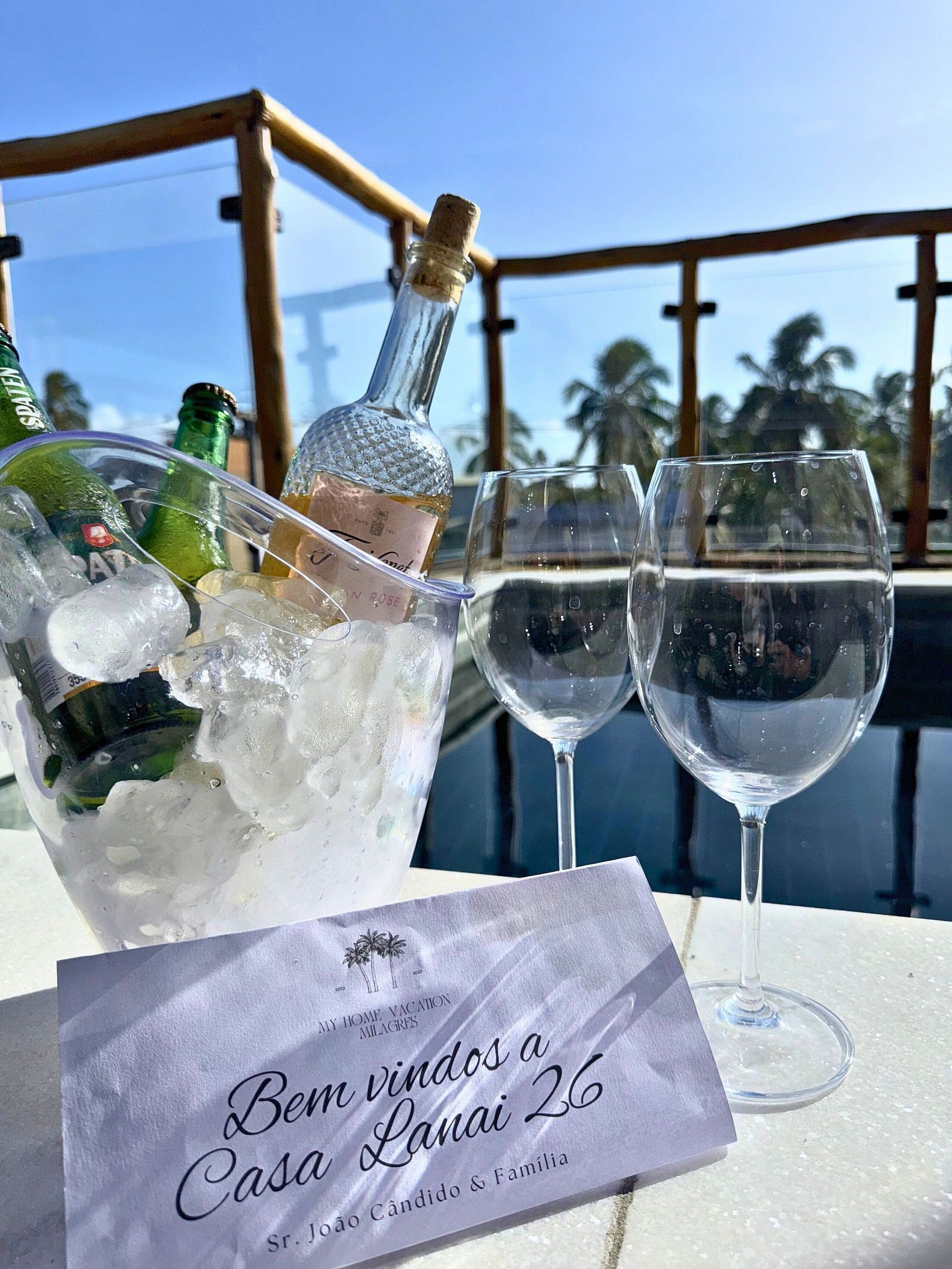 A bottle of champagne, two wine glasses, and a card on a table with a view of palm trees and a clear sky.