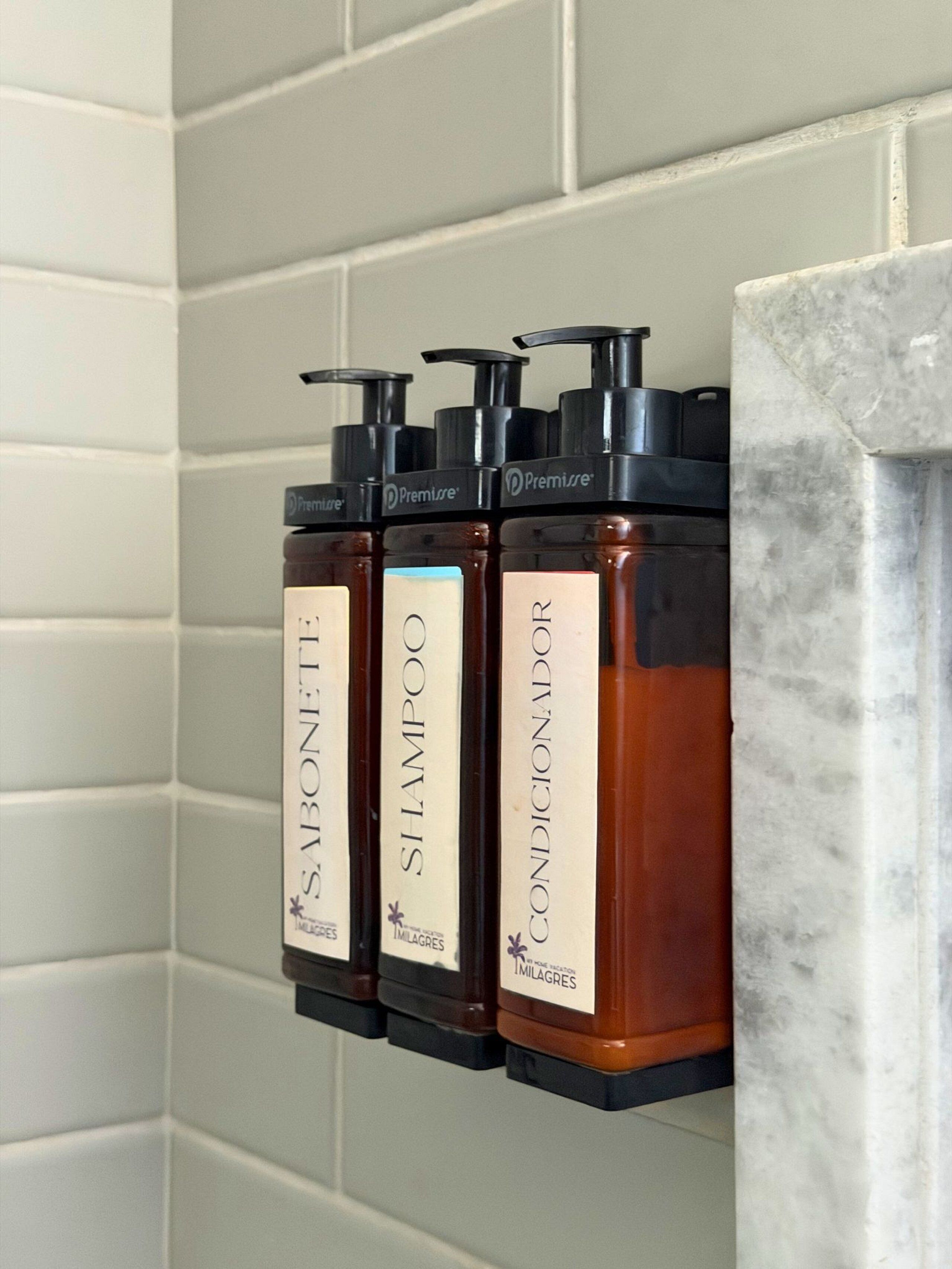 Three bottles of shampoo, conditioner, and soap on a tiled wall.