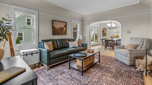 A living room with a green sofa, a grey armchair, a coffee table, and a dining area with a chandelier.
