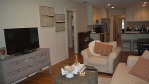 A living room with a television, a coffee table, and a sofa. There is a kitchen in the background with a refrigerator and a microwave. - 3-bedroom Cottage new LSU! (Baton Rouge)