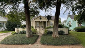 A two-story house with a front porch, surrounded by a picket fence and trimmed hedges. - 3-bedroom Cottage new LSU! (Baton Rouge)