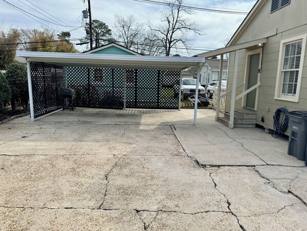 A covered patio area with a lattice fence, a concrete floor with visible cracks, and a house in the background. - 3-bedroom Cottage new LSU! (Baton Rouge)