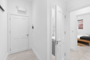 A hallway with white doors, a visible air conditioning unit, and a bed with a wooden frame in the background. - Modern 4 Beds Apt. in London with Parking + Garden (London)
