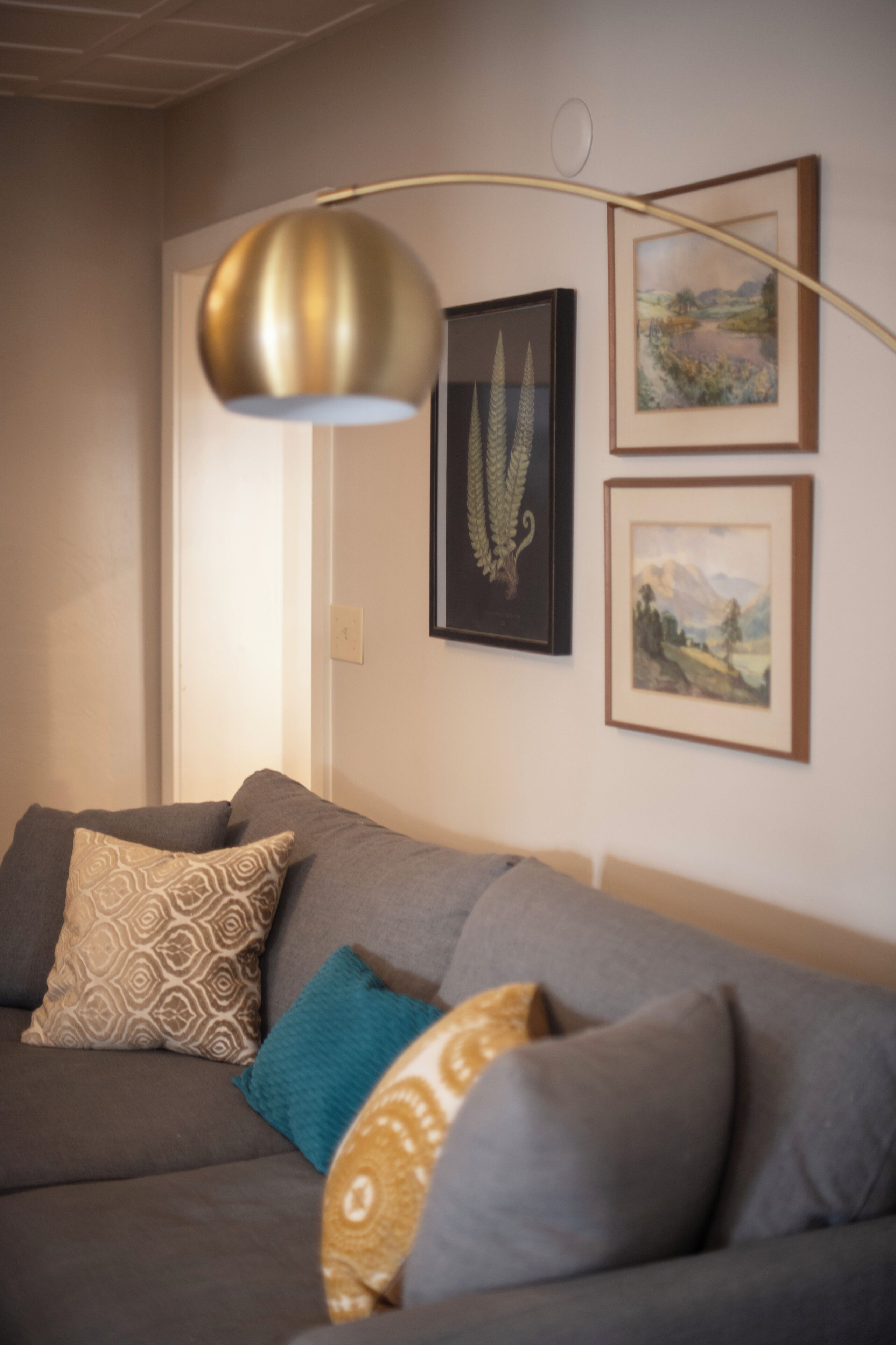 A living room with a grey sofa, patterned cushions, a gold pendant lamp, and framed landscape paintings on the wall.