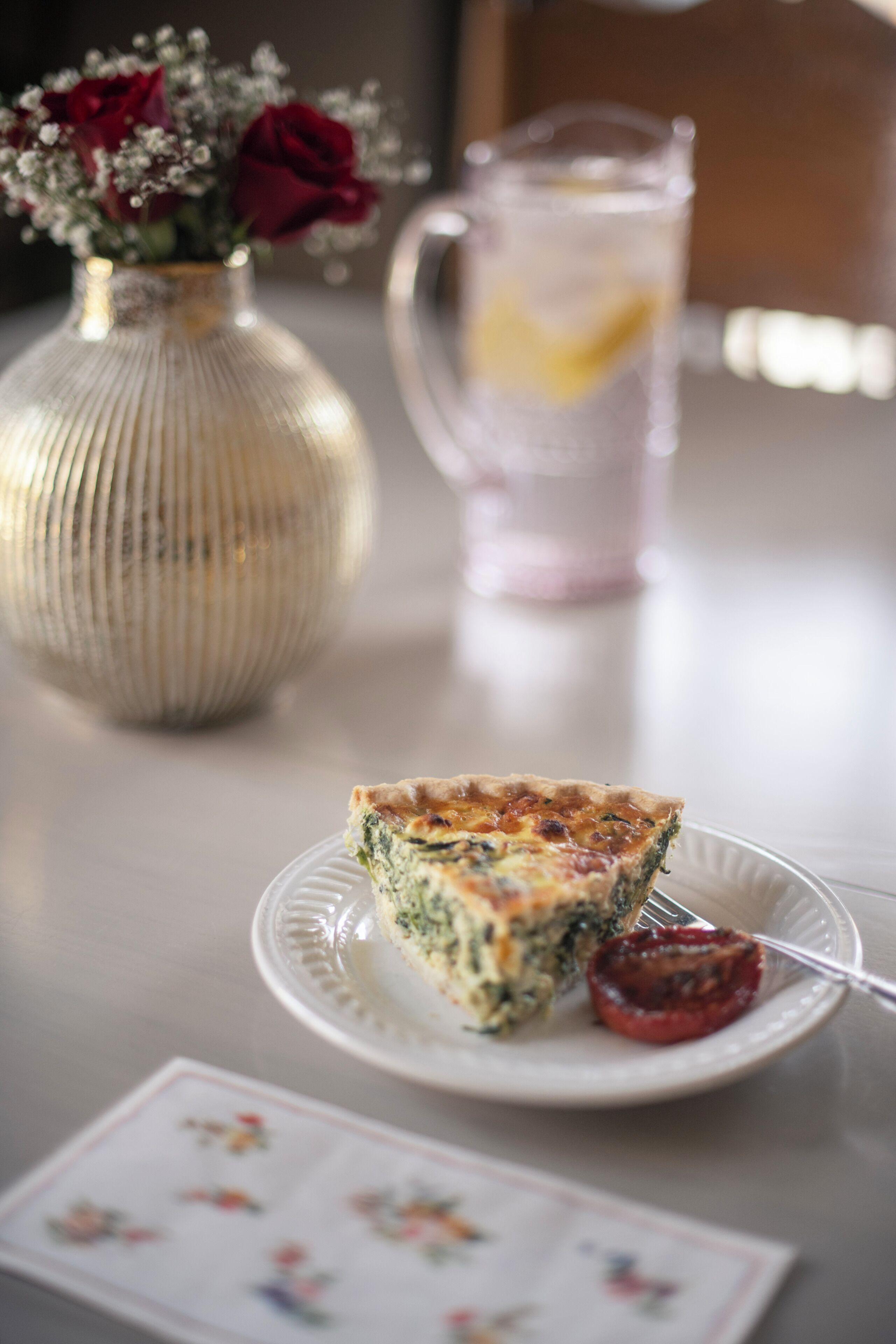 A slice of quiche on a plate with a fork, a pitcher of lemonade, and a vase with flowers on a table.