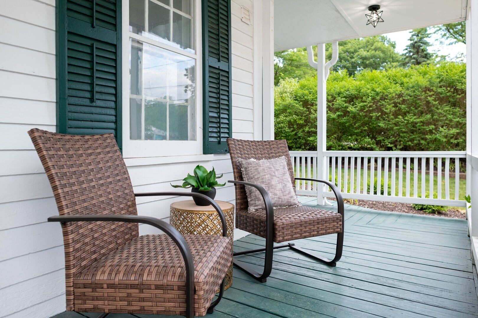 A porch with two wicker chairs, a small round table with a plant, and a white railing.