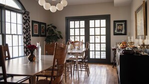 A dining area with a wooden table, chairs, a chandelier, and a large window with curtains.