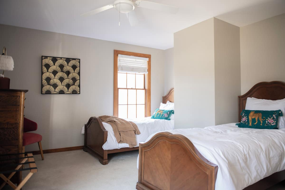 Une chambre avec deux lits, un ventilateur de plafond, une fenêtre avec des stores et une œuvre d’art accrochée au mur.