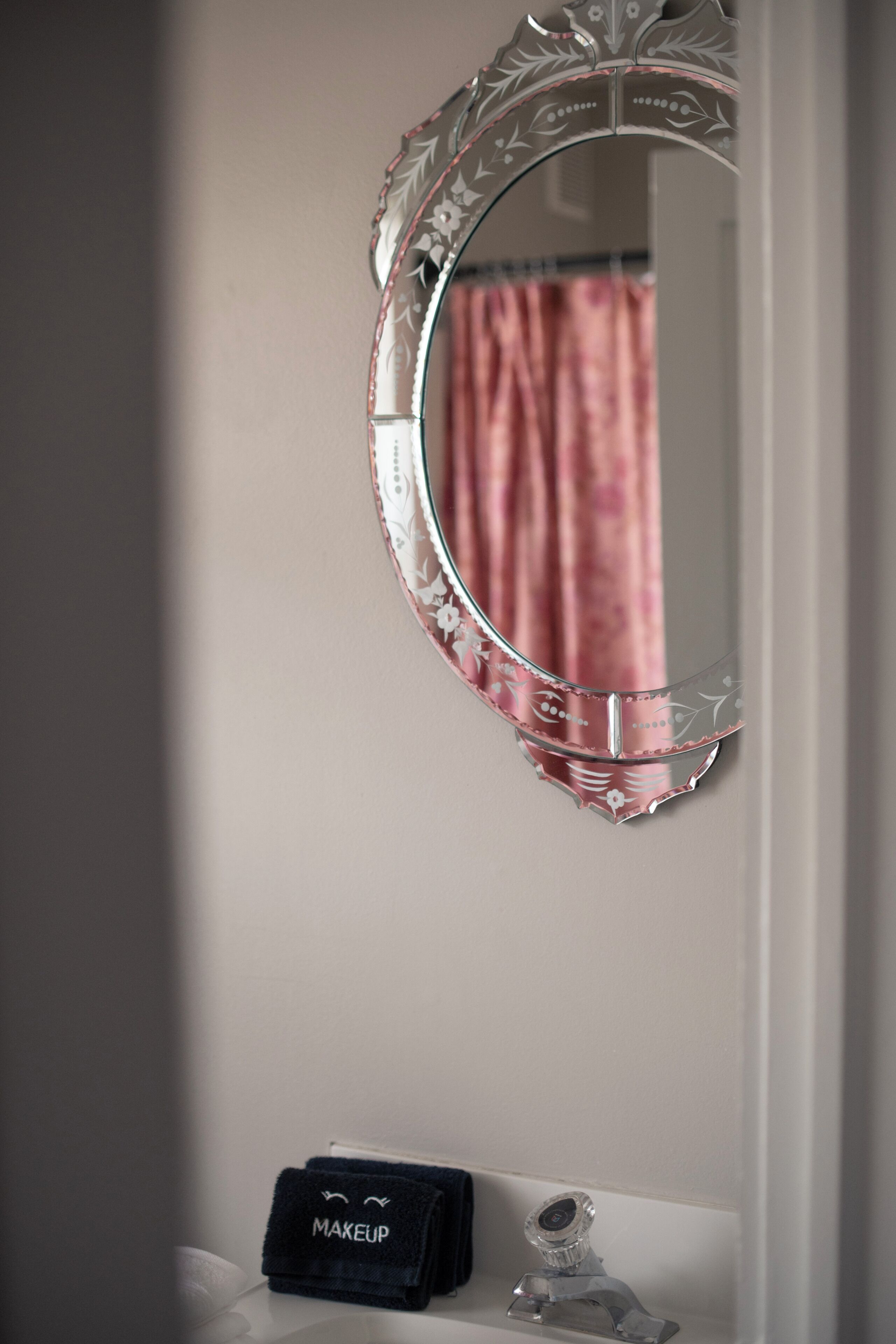 Et badeværelse med et rundt spejl, en vask, en makeuppose og en badeforhæng.