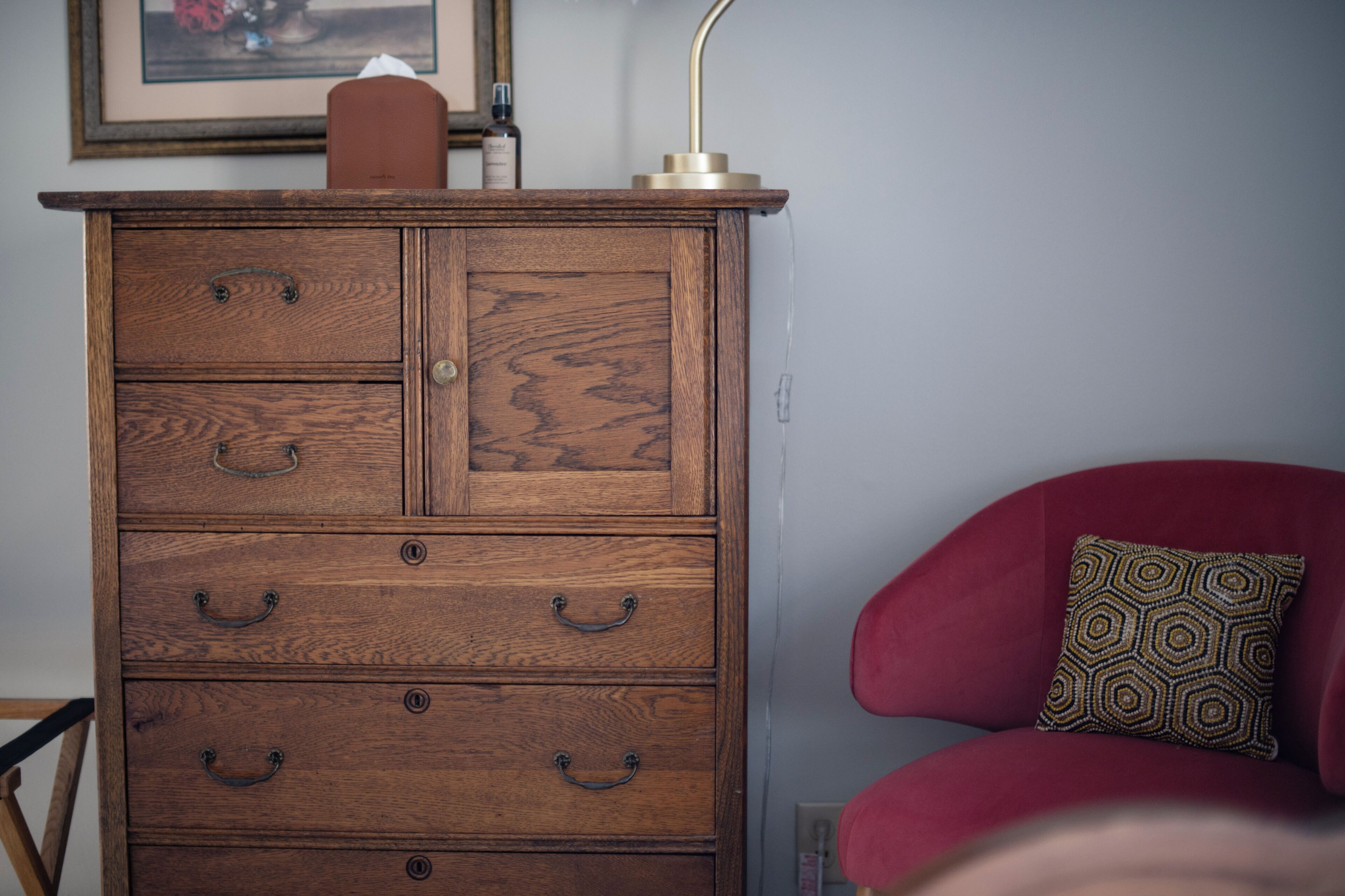 A wooden dresser with drawers and a small table on top, a red chair with a patterned cushion, and a framed picture on the wall.