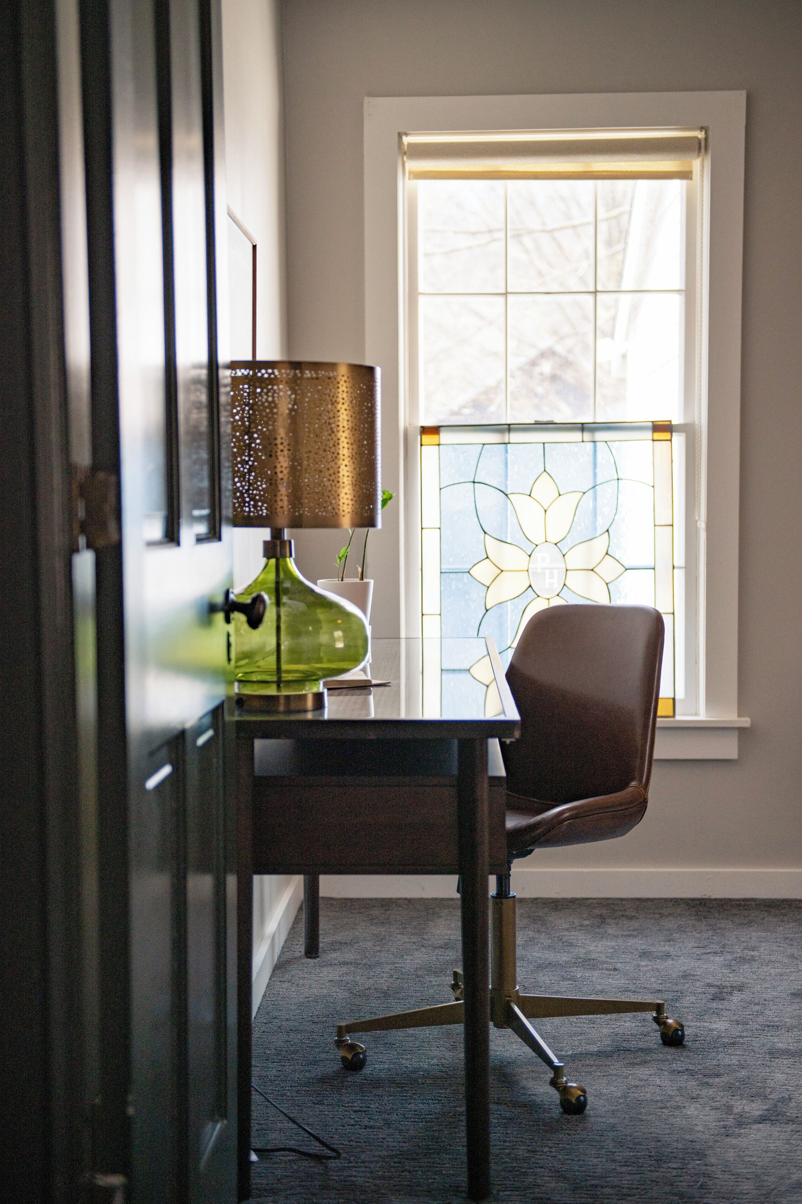A room with a desk, chair, lamp, and a window with a stained glass design.
