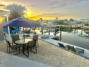 Une terrasse avec une table et des chaises, un parasol bleu, et une vue sur une marina avec des bateaux et un coucher de soleil.