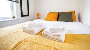 Uma cama bem arrumada com roupa de cama branca e laranja, um cobertor amarelo texturizado e duas toalhas brancas dobradas sobre a cama.