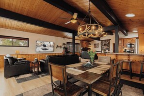 A spacious living area with a wooden ceiling, a dining table with chairs, a black leather sofa, and a kitchen in the background. - Peak & Quiet · Moonridge · Mountain View Escape · Spa, Yard, EV (Big Bear Lake)