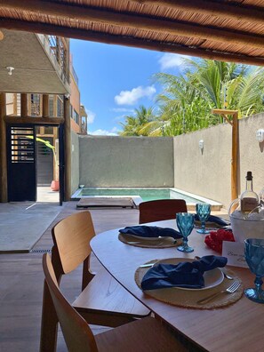 Uma área de refeições ao ar livre com uma mesa posta para dois, piscina ao fundo e vista para palmeiras.