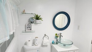 A bathroom with a glass sink, white fixtures, a round mirror, a toilet with a roll of toilet paper, a towel, and a small plant on a shelf.