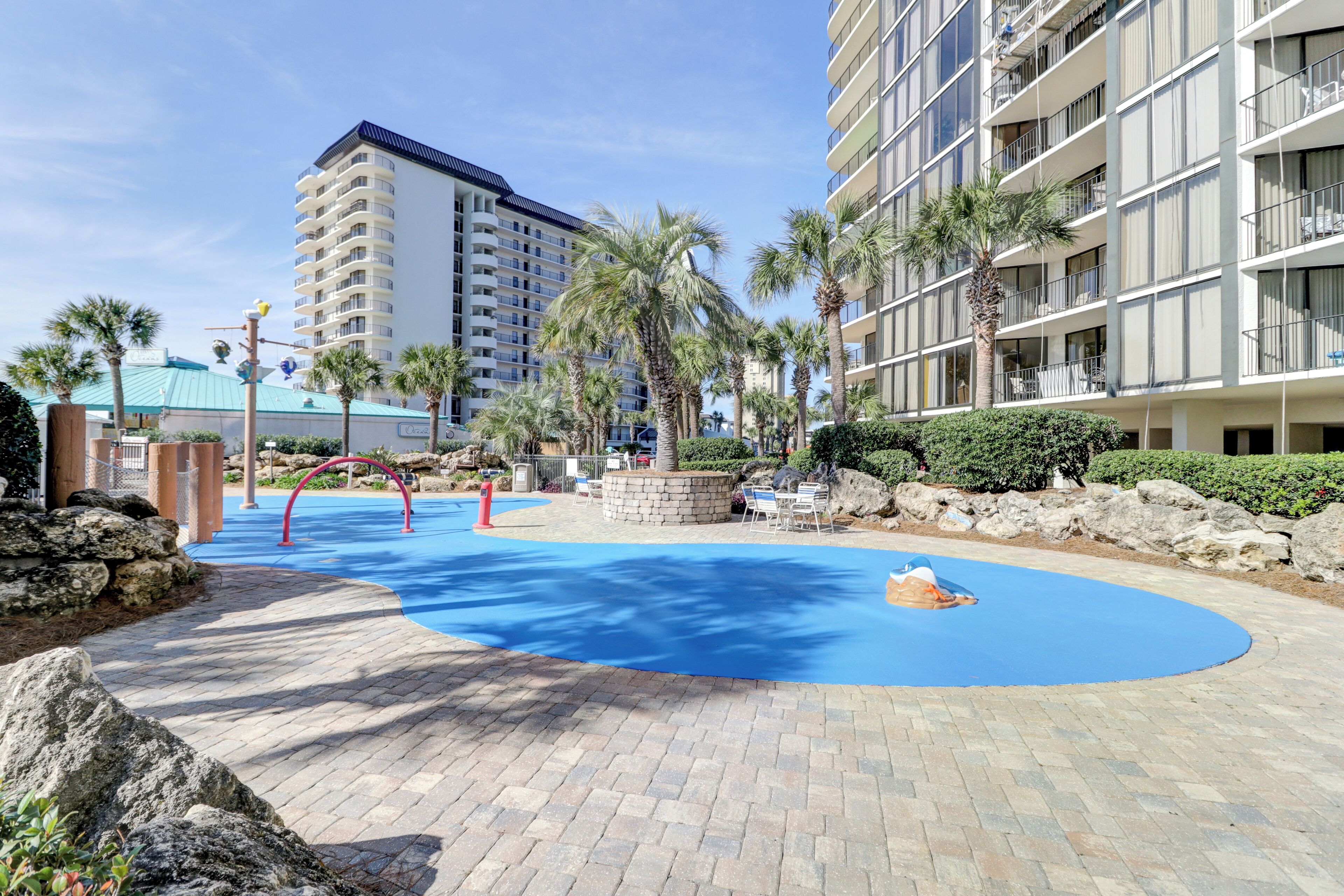 Un patio de hotel con piscina azul, palmeras y un edificio grande al fondo.