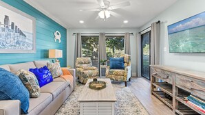 A living room with a teal accent wall, a ceiling fan, a large window with curtains, a sofa with patterned cushions, a coffee table, and a bookshelf. - Billie Jeans Bungalow! Cozy Edgewater Condo w/ Resort Amenities, Sleeps 6 (Panama City Beach)