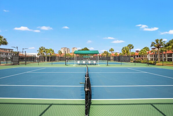 An outdoor tennis court with a net, surrounded by palm trees and buildings. - Billie Jeans Bungalow! Cozy Edgewater Condo w/ Resort Amenities, Sleeps 6 (Panama City Beach)