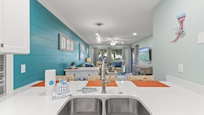 A modern kitchen with a double sink, a dining area with orange placemats, and a living room with a TV and a ceiling fan. - Billie Jeans Bungalow! Cozy Edgewater Condo w/ Resort Amenities, Sleeps 6 (Panama City Beach)