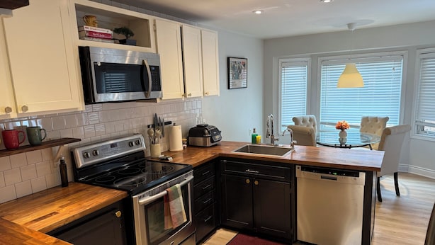 A modern kitchen with white cabinets, a built-in microwave, a stove, and a dishwasher. There's a wooden countertop, a red doormat, and a window with blinds. - Elegant 4BR Home | Private Backyard & Trail Access (Waterloo)