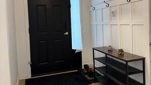 A hallway with a black door, a black mat, a wooden floor, a black metal shelf with potted plants, and a hanging light fixture. - Elegant 4BR Home | Private Backyard & Trail Access (Waterloo)