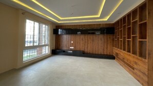 A spacious room with a large window, wooden shelving, and a flat-screen TV mounted on the wall.