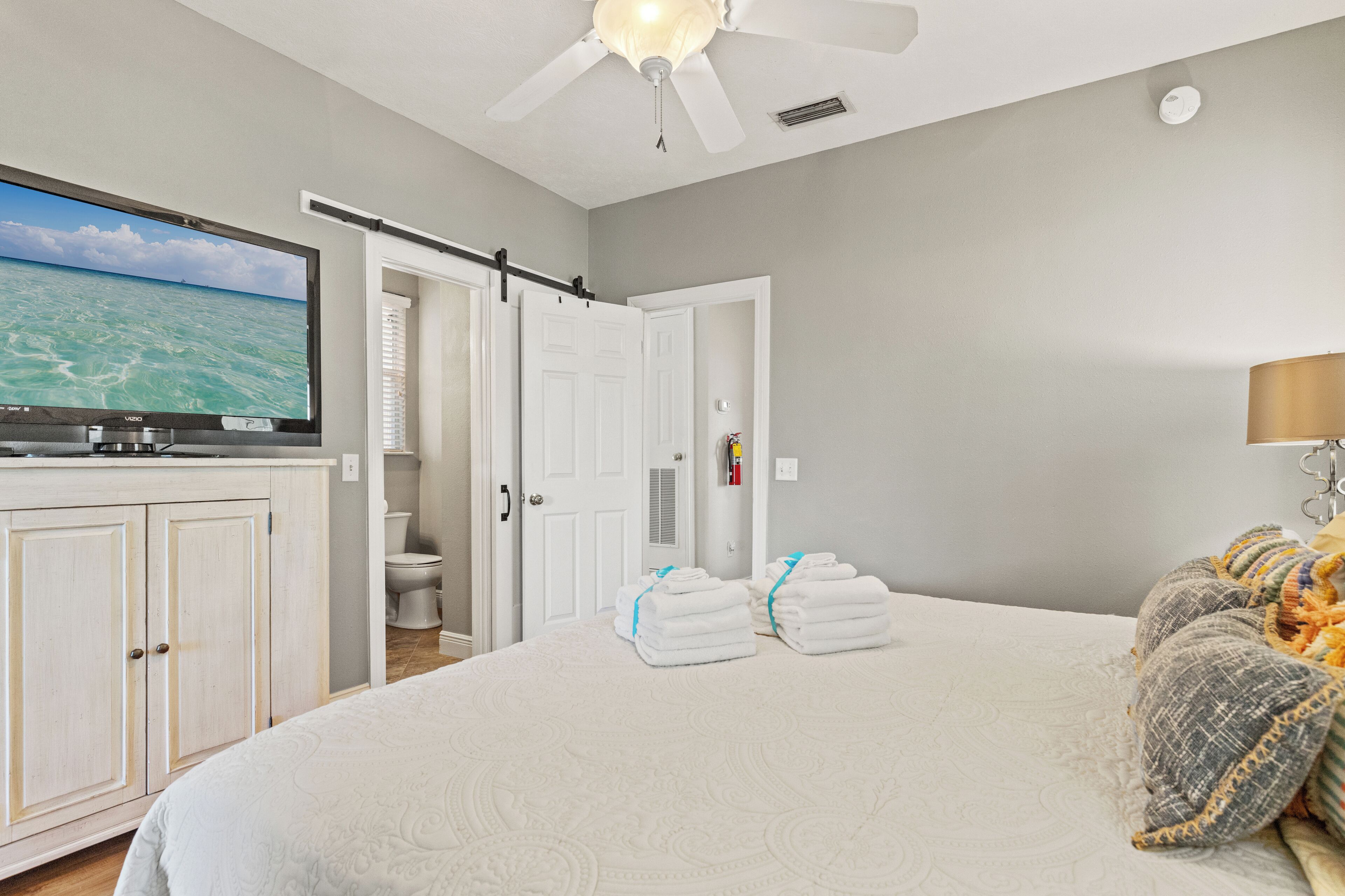 A bedroom with a bed, a TV, a ceiling fan, a bathroom, and a door.