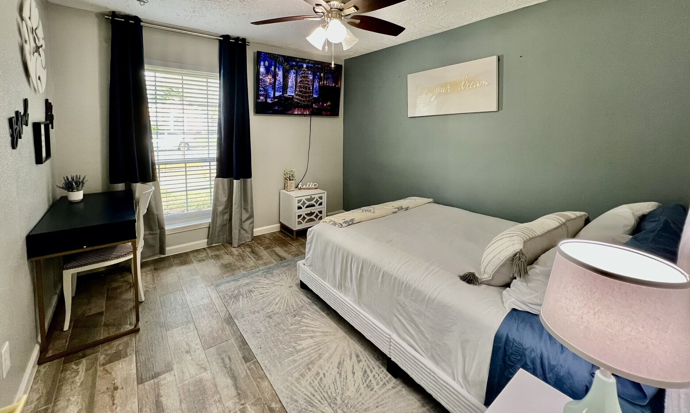 A bedroom with a bed, a desk, a ceiling fan, a TV, and a motivational sign on the wall.