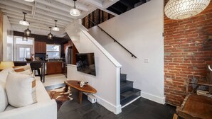 A modern living space with a staircase leading to a loft area, a dining area with a wooden table, and a kitchen in the background. - Romantic Designer Loft | Walk to Butler St! (Pittsburgh)