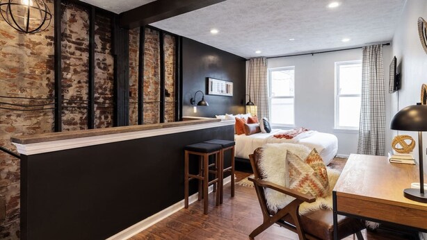 A modern hotel room with a brick accent wall, a large bed, a desk, and a bar area. - Romantic Designer Loft | Walk to Butler St! (Pittsburgh)