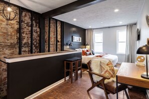 A modern hotel room with a brick accent wall, a large bed, a desk, and a bar area. - Romantic Designer Loft | Walk to Butler St! (Pittsburgh)