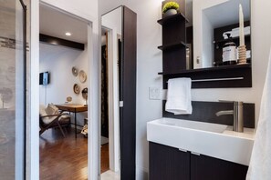 A modern bathroom with a white sink, dark wood vanity, and a mirror above the sink. There is a towel hanging on the wall and a plant on the shelf. - Romantic Designer Loft | Walk to Butler St! (Pittsburgh)