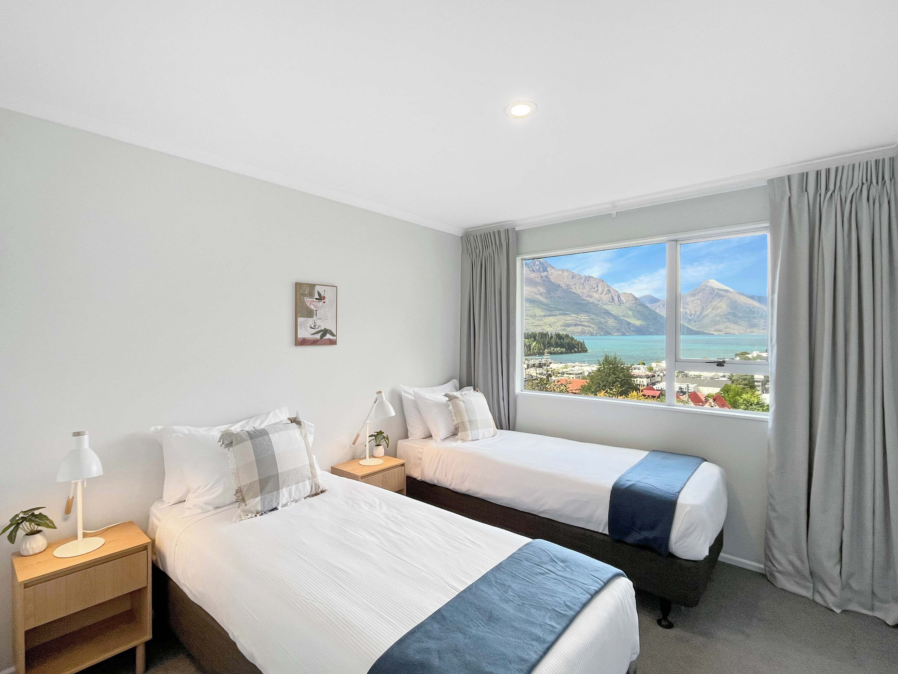 Quarto de hotel com duas camas, mesa de cabeceira com abajur, pequena mesa com planta e janela com vista para a montanha.