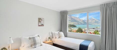 Quarto de hotel com duas camas, mesa de cabeceira com abajur, pequena mesa com planta e janela com vista para a montanha.