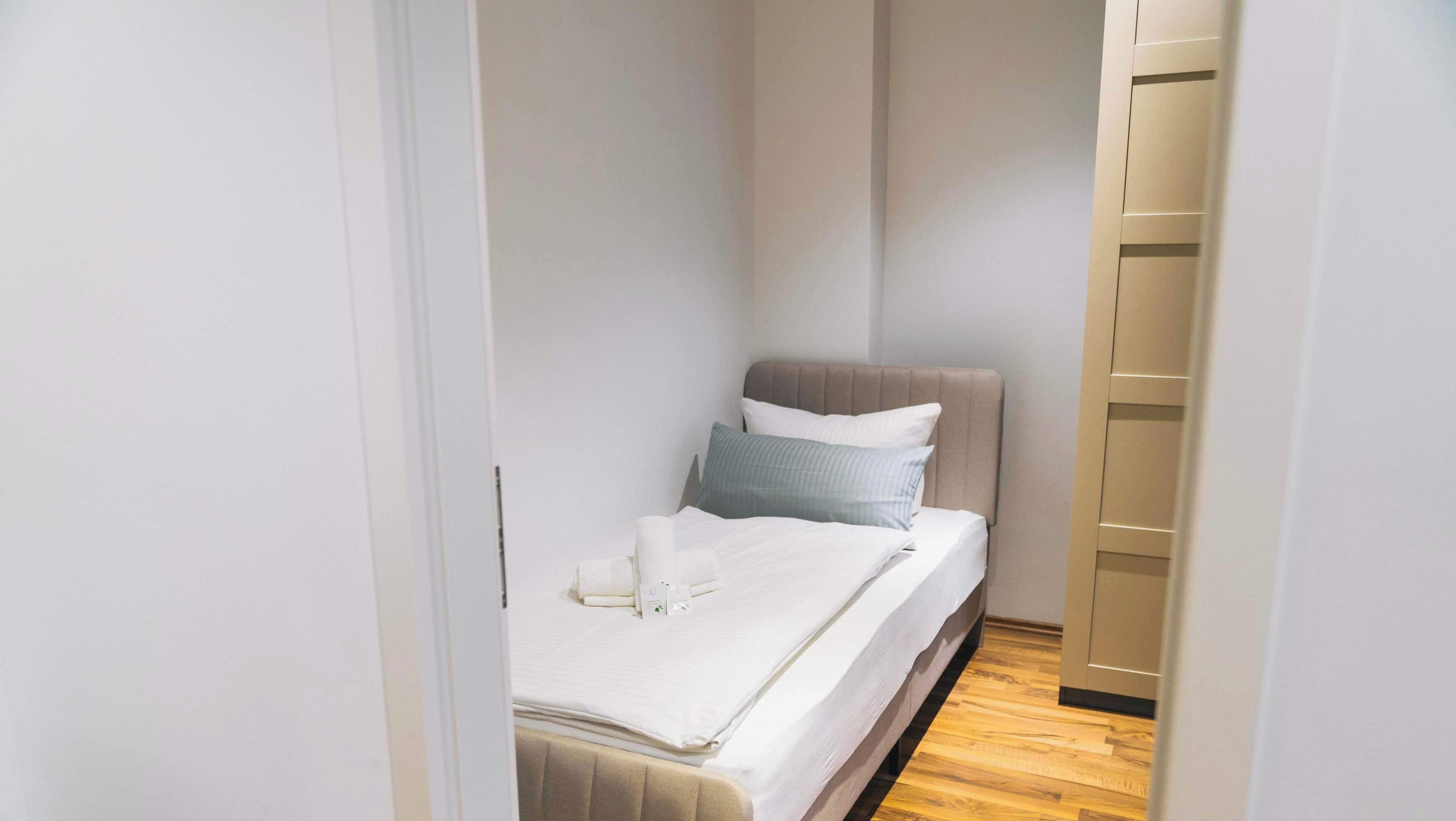 A narrow room with a bed, a bedside table, and a wooden floor.