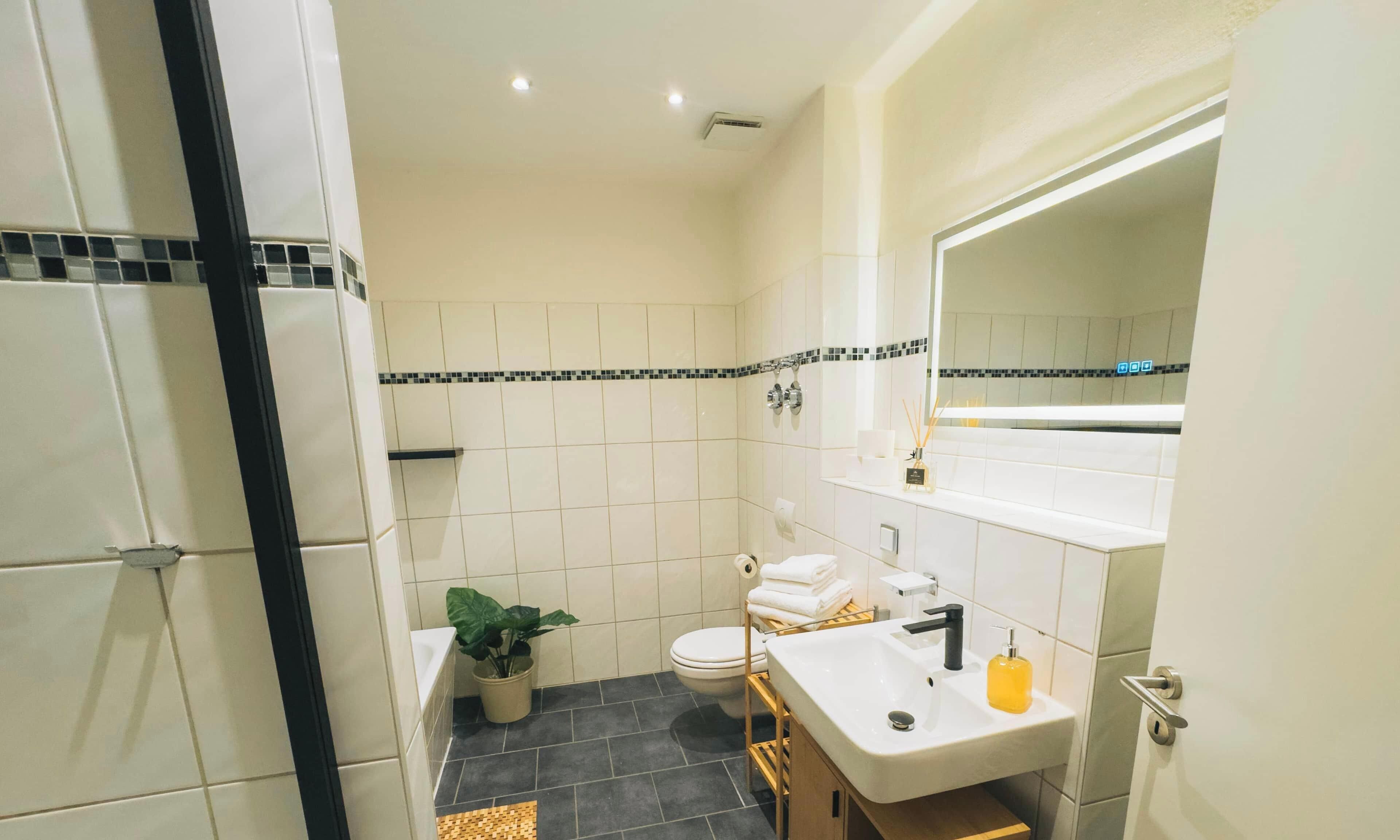 Un baño con azulejos blancos, un borde blanco y negro, un lavabo con dispensador de jabón amarillo, un inodoro y una planta en maceta.