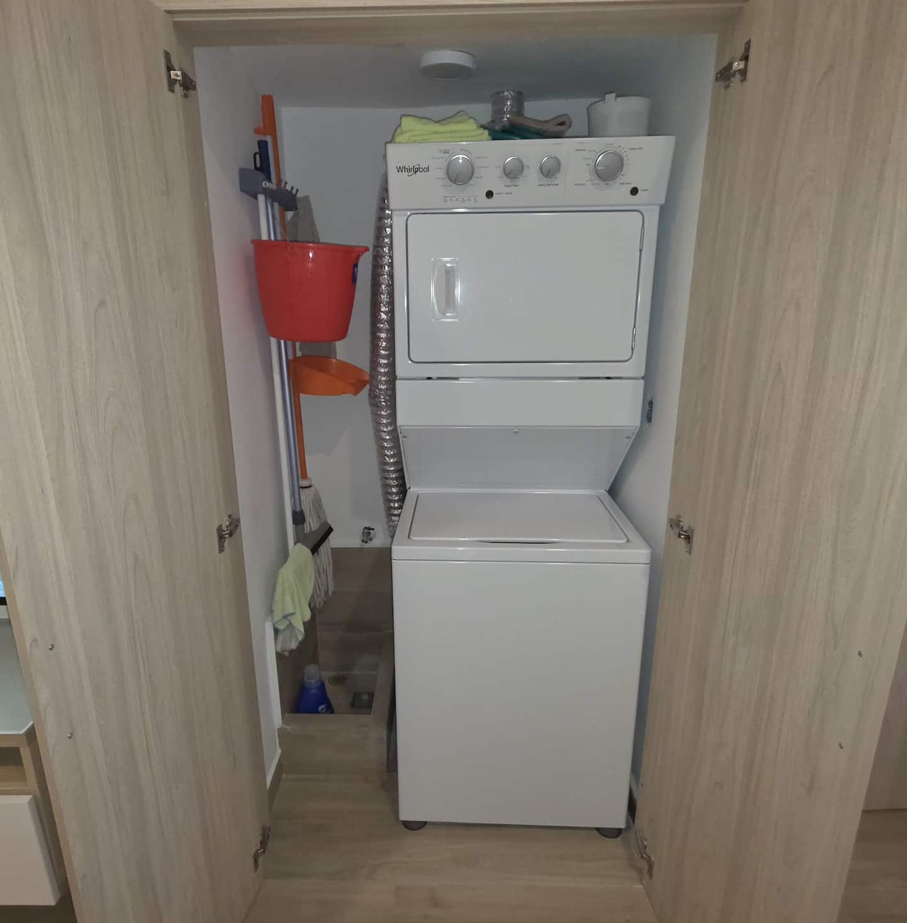 A compact laundry room with a stacked washer and dryer, a red mop bucket, and a mop on a pole.