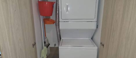Un lavadero pequeño con lavadora y secadora apiladas, un cubo rojo para fregar y un trapeador.
