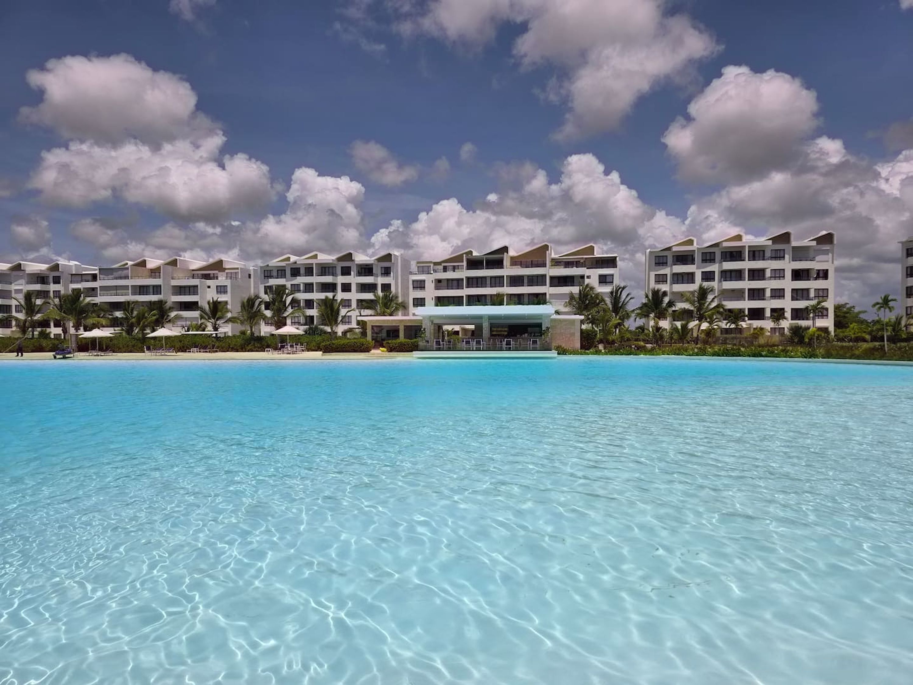 Un resort con una piscina amplia, varias edificaciones blancas y palmeras.
