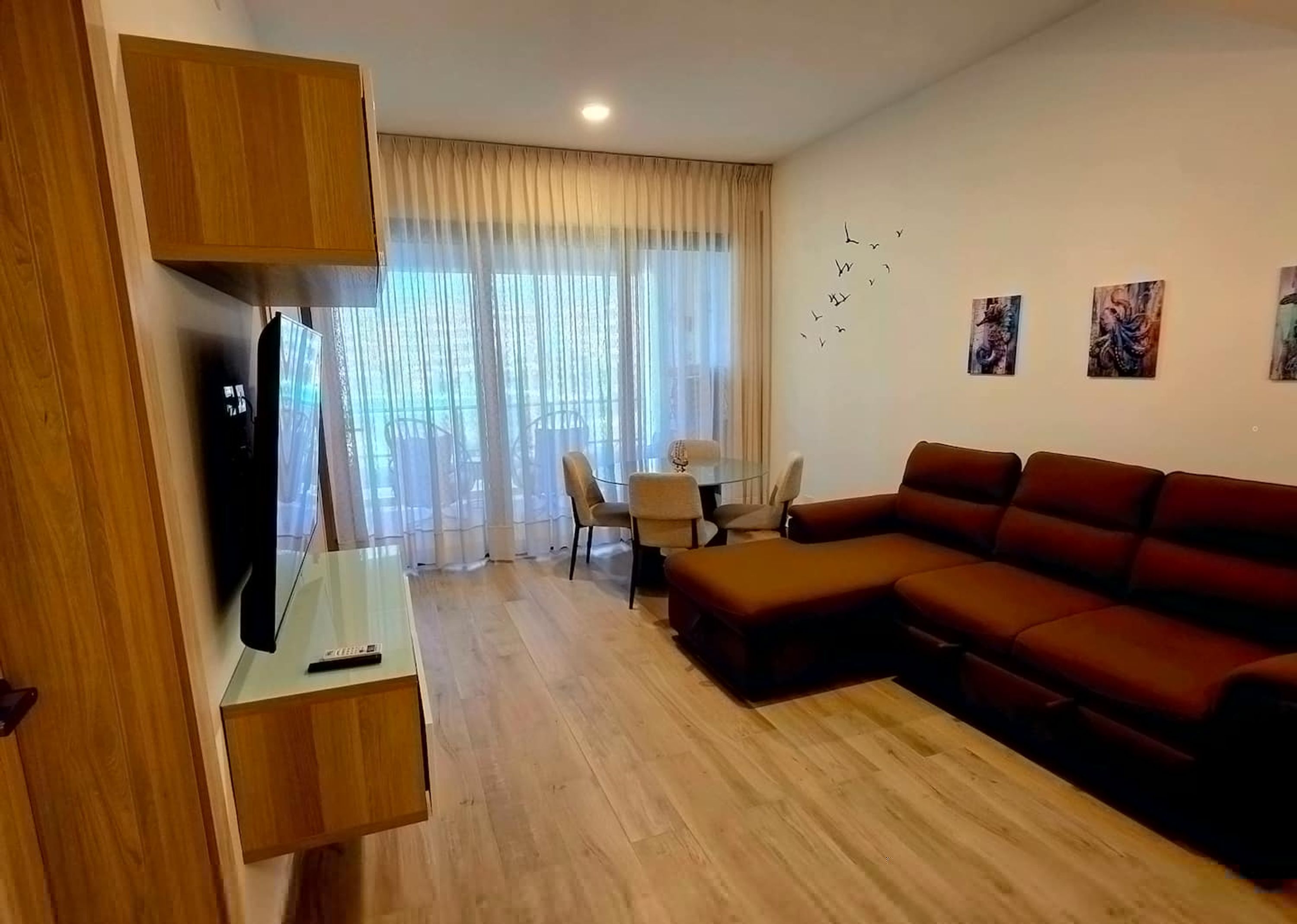 A modern living room with a sectional sofa, a TV, a dining table with chairs, and a large window with sheer curtains.
